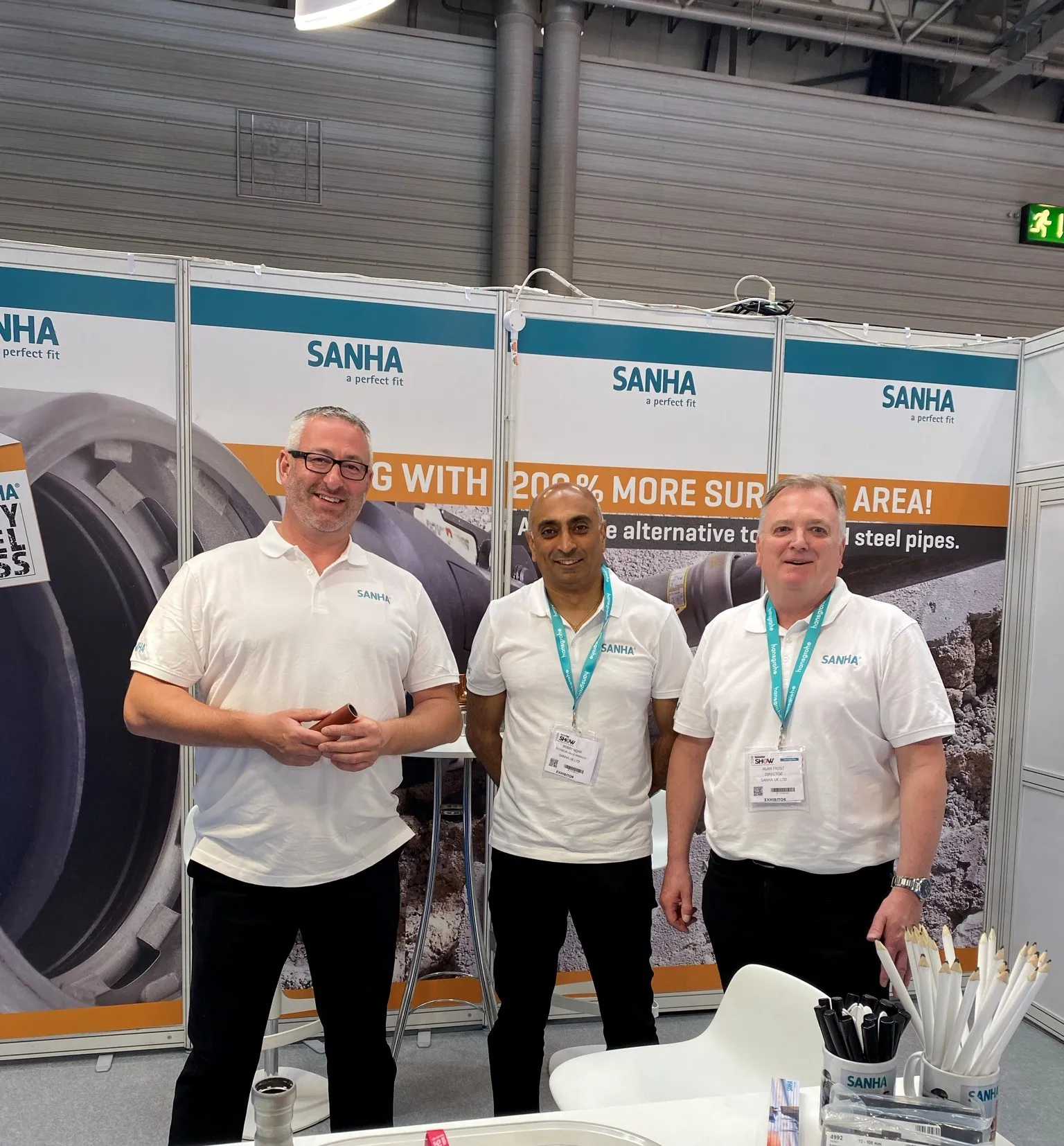 Three employees of SANHA® UK with white shirts are centered in front of a convention wall